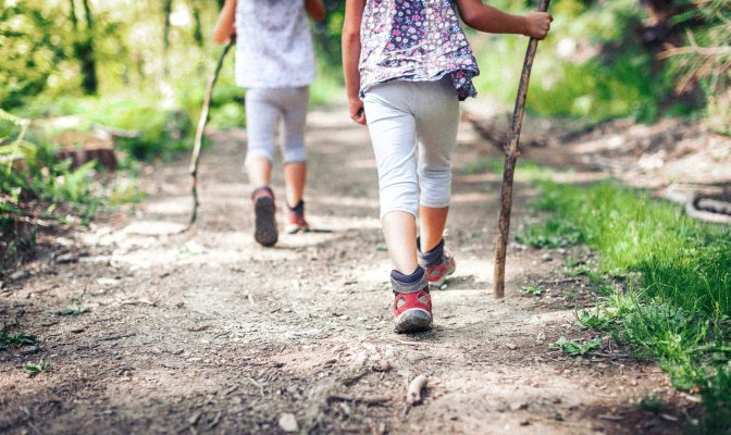 Children hiking in mountains or forest with sport hiking shoes. Girls are walking trough forest path wearing mountain boots and walking sticks. Frog perspective with focus on the shoes.